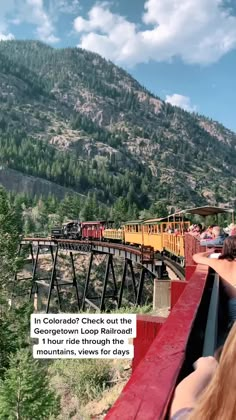 a train traveling over a bridge next to a lush green forest filled mountain side under a blue sky with white clouds