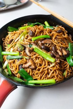 a wok filled with noodles, mushrooms and green onions next to chopsticks