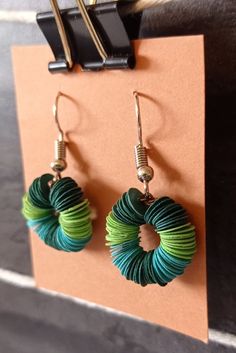 two pairs of green and blue earrings hanging from hooks on a brown paper clipboard