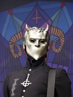 a man wearing a white mask and black outfit with horns on his head, standing in front of a stained glass window