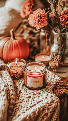 two candles sitting on top of a blanket next to flowers and a vase with pumpkins