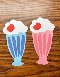 two cupcakes cut out from paper on top of a wooden table, one is blue and the other is pink