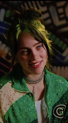 a woman with green hair and piercings smiles at the camera while wearing a green jacket