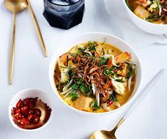 two bowls filled with food next to gold spoons on a white tablecloth and utensils