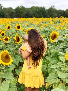 a woman in a yellow dress holding a baby standing in a field of sunflowers