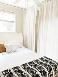 a white bed sitting under a window next to a white curtain covered wall in a bedroom