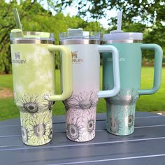 three different colored coffee mugs sitting on top of a table in front of trees