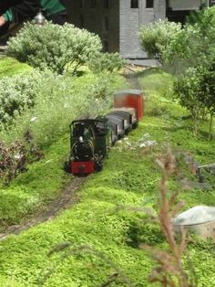 a small train traveling through a lush green forest next to a tall brick building on top of a hill