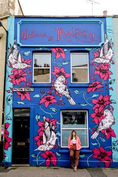 a woman standing in front of a blue building with pink flowers painted on the side