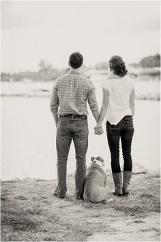 a man and woman holding hands while standing next to a dog on the beach with water in the background