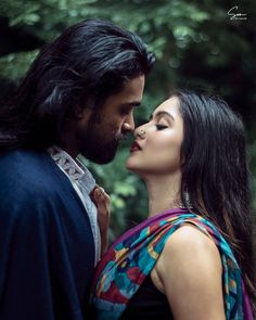 a man and woman standing next to each other in front of trees with their noses close together