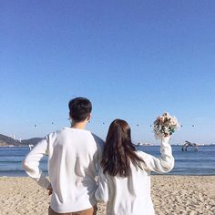 a man and woman standing on top of a sandy beach next to the ocean holding flowers