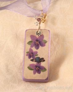 a necklace with purple flowers is hanging on a white fabric background and has a ribbon around it