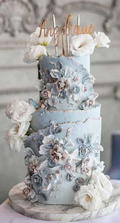 a blue wedding cake with white flowers and gold lettering on top is sitting on a table