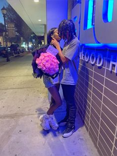 two people standing next to each other kissing and holding flowers in front of a building