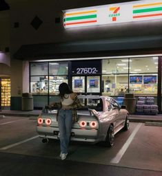 a woman leaning on the hood of a silver car in front of a convenience store