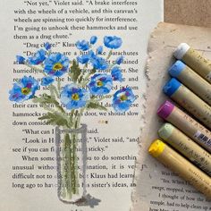 an open book with crayons next to it and some flowers in a vase