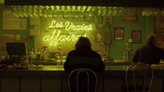 two people sitting at a bar in front of a neon sign that reads les vivres affair