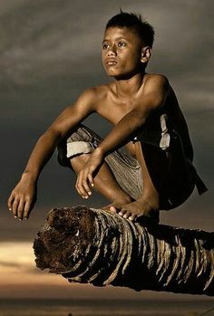 a young boy sitting on top of a tree branch in front of a cloudy sky