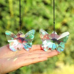 two small crystal butterflies hanging from strings in someone's hand with trees in the background