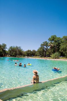 people are swimming in a large pool surrounded by trees and blue sky, with one person sitting on the edge