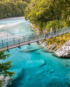 two people walking across a bridge over a blue river in the mountains near a forest