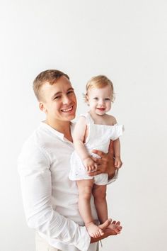 a man holding a baby in his right hand and smiling at the camera while standing against a white wall