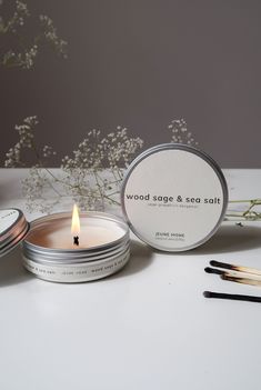 two tins of wood sage and sea salt candles on a table with flowers in the background