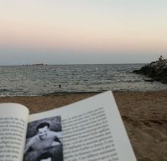 an open book sitting on top of a sandy beach next to the ocean with people swimming in the water