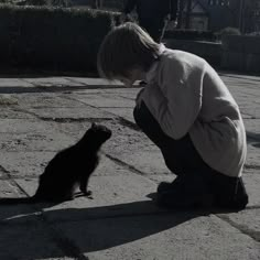 a young boy kneeling down next to a black cat on the ground with his head in his hands