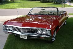 an old red convertible car parked on the grass in front of a driveway and house