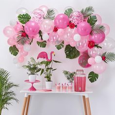 a table topped with pink and white balloons next to palm leaves, plants and flamingos