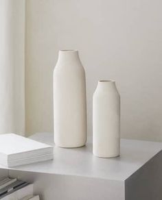 two white vases sitting on top of a table next to books and a window