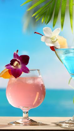 two glasses filled with different colored drinks next to each other on a table near the ocean