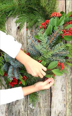 two hands are holding evergreen leaves and berries on a wooden background with text overlay that reads how to make an evergreen wreath