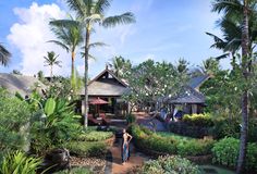 a woman standing in the middle of a lush green area with palm trees and buildings