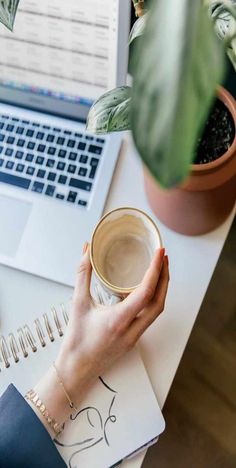 a woman holding a cup of coffee while sitting at a desk with a laptop and potted plant