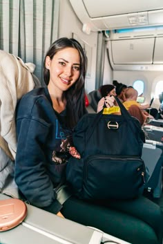 a woman sitting on an airplane holding a black backpack and smiling at the camera with other passengers in the background