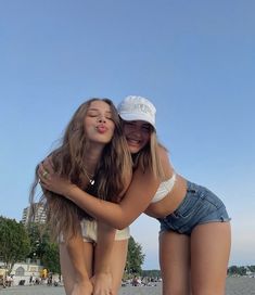 two girls are hugging on the beach while one girl is wearing a white hat and blue jean shorts