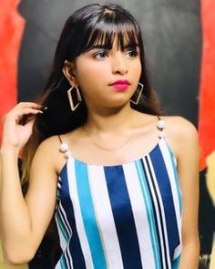 a woman with long black hair wearing large earrings and a striped top standing in front of a painting