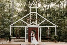 a bride and groom standing in front of a white house with red doors, surrounded by greenery