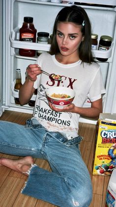 a woman sitting on the floor in front of an open refrigerator holding a bowl of cereal