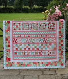 a red and green quilt sitting on top of a wooden fence next to pink flowers