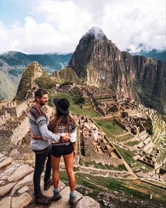 a man and woman standing on top of a mountain looking at an ancient city in the distance