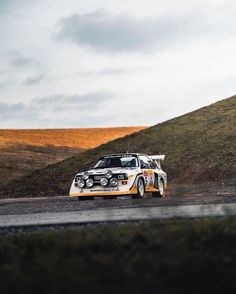 an old race car driving down the road in front of a hill with grass on it