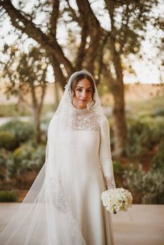 a woman in a wedding dress and veil holding a bouquet standing next to a tree