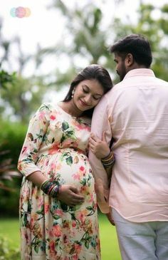 a pregnant woman standing next to a man in a pink shirt and floral print dress