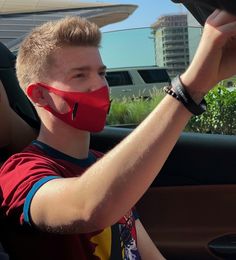 a young man wearing a red face mask while sitting in a car with his hand on the steering wheel