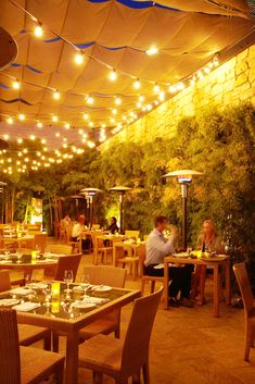 people are sitting at tables in an outdoor dining area with lights strung over the ceiling