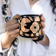 a woman is holding a black and orange coffee mug with flowers on it, while wearing a white shirt